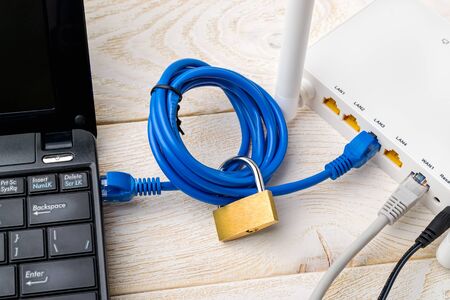 Padlock on a blue network cable connecting laptop and white WiFi router on a white wooden  table....