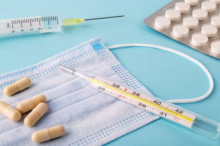 Disposable surgical mask, thermometer, pills and syringe on a blue background. Medical mask reduces the risk of infection. Airborne disease protection concept.の写真素材