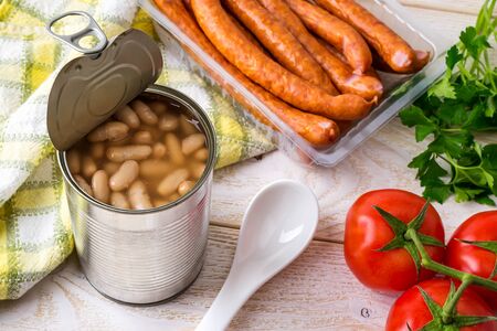 Canned backed beans in an open tin can with pull tab near thin sausages, tomatoes and ceramic spoon on a wood table. Bean is source of vegetable protein and ingredient for vegetarian food. Top view.の写真素材