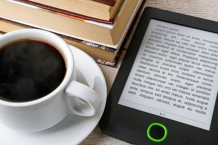 Close-up of e-book reader near cup of hot coffee and stack of ordinary paper books. Copy space on e-book display. E-reading for pleasure and education. Front view.の写真素材