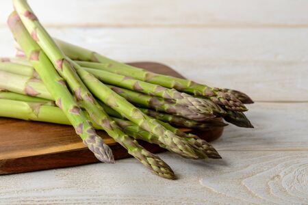 Raw fresh green asparagus stalks on a brown cutting board over a white wood kitchen table. Ingredient for healhy eating, vegetarian and raw food diet. Front view.の写真素材