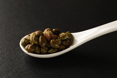 Pickled capers in a white chinese porcelain spoon on a black stone serving board. Sauces and seasonings for meat, fish and vegetables. Front view.の写真素材