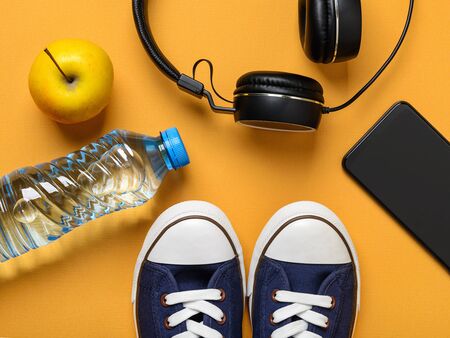 Blue sneakers or gumshoes with white shoe laces, smartphone, headphones, ripe apple and bottle of drinking water on a textured yellow background. Shoes for fitness and sport. Flat lay.の写真素材
