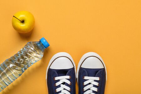 Classic blue sneakers or gumshoes with white shoe laces, ripe apple and bottle of drinking water on a textured bright yellow background. Comfortable shoes for fitness and sport. Copy space. Flat lay.の写真素材