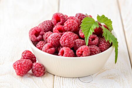 Whole ripe sweet raspberries and green leaf in a beige ceramic bowl on a white wood table. Tasty...