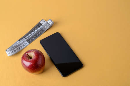 Measuring tape with centimeters, smartphone and ripe red apple on a textured yellow background. Concept of healthy eating, dieting, weight loss and body slim. Copy space.の写真素材