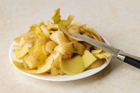 Heap of fresh potato peels and small vegetable knife on a white plate ober kitchen table. Cook boiled potatoes at home. Household biodegradable waste. Top view.の写真素材