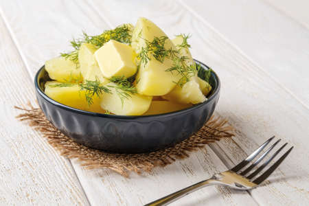 Delicious freshly boiled potatoes with melting butter and dill in a ceramic bowl on a white wooden table. Cook at home delicious vegetable dish. Healthy eating. Front view.の写真素材
