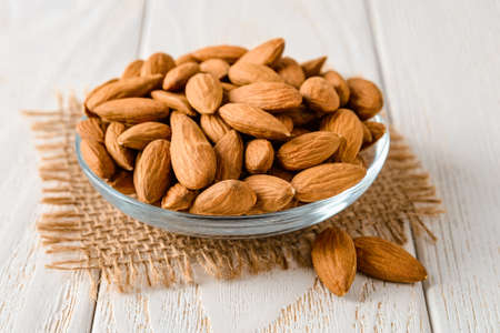 Tasty raw almonds in a glass saucer on a white wood table. Unroasted nuts as antioxidant and protein source for ketogenic diet and vegetarianism. Healthy eating. Front view.の写真素材