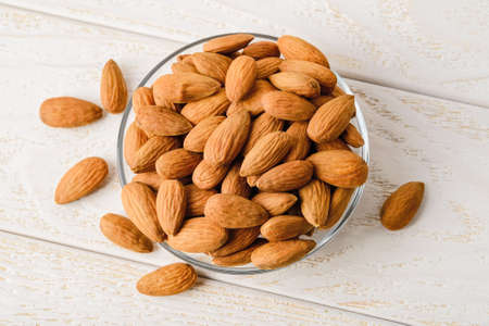 Heap of raw almonds in a glass saucer on a white wood table. Unroasted nuts as antioxidant and protein source for ketogenic diet and vegetarianism. Healthy eating. Top view.の写真素材