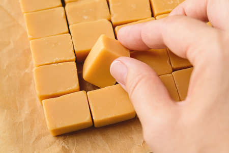 Woman hand takes one caramel vanilla fudge from a brown parchment paper. Fresh tasty candies made of...