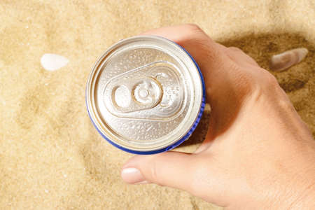 Woman hand takes cold beer or soda can with water drops from sea sand on a sunny day. Drinking on...