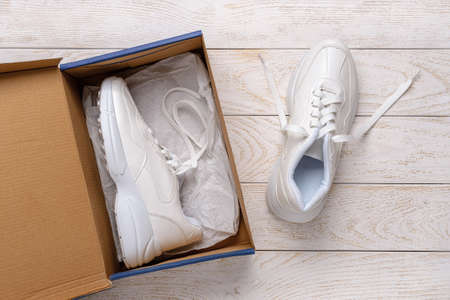 One white chunky sole sneakers in a brown cardboard box, the other on the white wood floor. Open box with new comfortable shoes for active lifestyle, fitness and sports. Top view.の写真素材