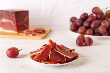 Sliced jamon on a plate against large jamon piece on a cutting board and bunch of pink grapes over...