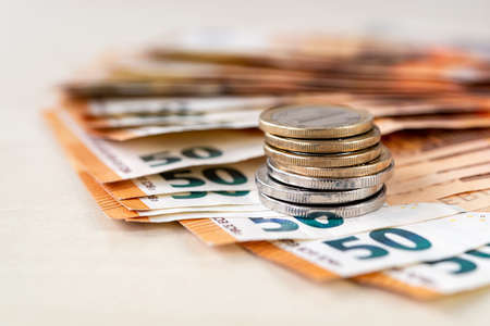 Stack of coins on 50 euro banknotes folded in a fan on a beige tabletop. Wealth, cash savings, accounting of income and expenses concepts. Shallow depth of field. Front view.の写真素材