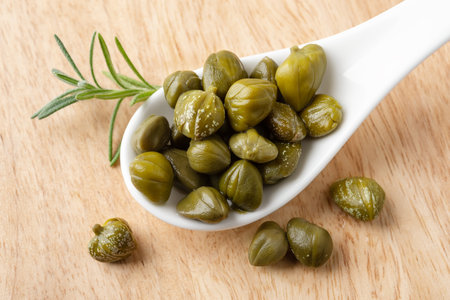 Pickled capers in a white porcelain spoon on a brown wooden cutting board. Marinated buds of caper...