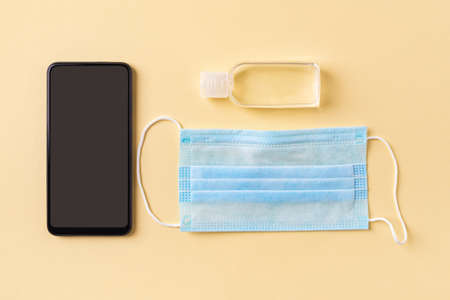Disposable medical face mask, hand sanitizer in a small bottle and smartphone on a yellow...