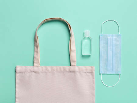 Eco-friendly cotton grocery bag, hand sanitizer in a small bottle and medical face mask on a green...