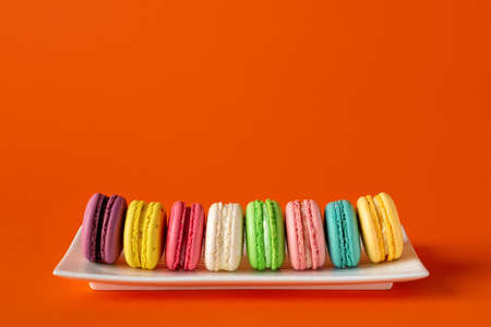 Multicolored macaroons in a row on a white rectangular plate against bright red background. Mockup of greeting card with colorful macaron cakes. Delicious french almond meringue cookies. Copy space. Front view.の写真素材