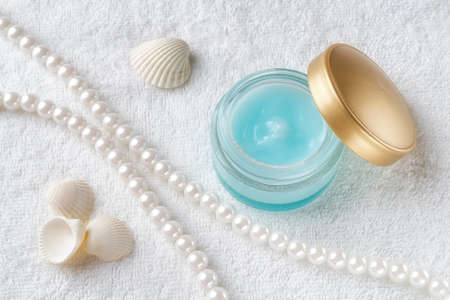 Moisturizing facial gel in an open jar with golden lid, pearl beeds and few sea shell on a white towel background. Glass container of hydrating face cream. Beauty, skincare and cosmetology. Health care. Top view.の写真素材