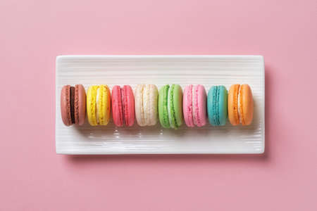 Multicolored macaroons on a white rectangular ceramic plate against pastel pink background. Sweet...