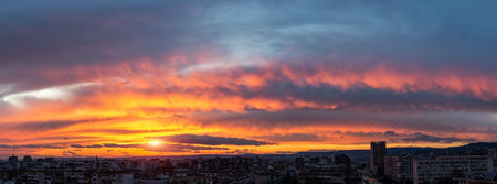 Dramatic sunset fire in the sky over a small city. Evening sun is low on the horizon. Panoramic landscape with urban skyline. Vivid red orange and dark blue sky during sundown. Scenic skyscape.の写真素材