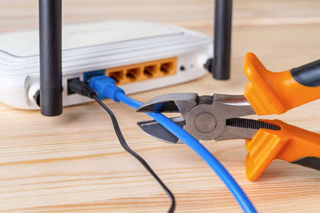 Wire snippers cutting the blue network cable connected to white Wi Fi wireless router on a wooden desk. Prohibit and restrict access to the internet, limit internet connection and internet censorship concepts. Close-up.の写真素材