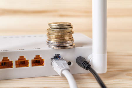 Stack of coins on the white Wi Fi wireless router with connected cables on a wooden desk. Monthly payment of internet, pay bills and earn money online concepts. Internet hardware and e-commerce. Close-up.の写真素材