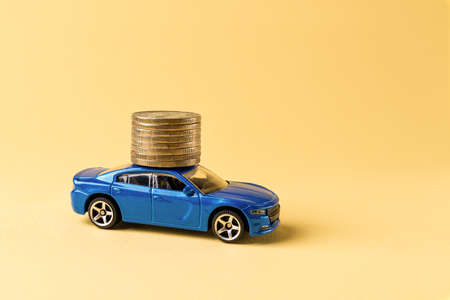 Blue toy car is carrying a stack of coin against yellow background. Saving money for a car. Car investment. Cost of car buying, service, repair, insurance and renting concepts. Copy space. Front view.の写真素材