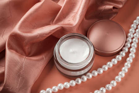 Beauty skin care cream in an open glass jar and white pearl beads near pale burgundy folded silk...