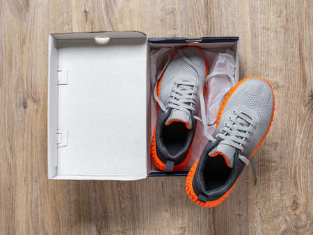 New sneakers in the open box on the floor. Pair of gray mesh fabric trainers with grooved orange sole in a cardboard box. Modern textile sneakers for sport and active lifestyle. Open new shoes box top view.の写真素材