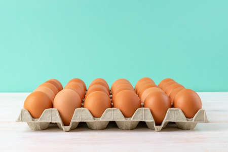 Brown chicken eggs in the cardboard tray on a white wooden table against turquoise background. Raw...