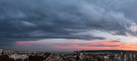 Stormy gray clouds is coming to seaside city at sunset. Panorama of dramatic overcast sky at evening. Dark stratus clouds hang over Varna, Bulgaria. Weather change concept. Storm is coming wide landscape.の写真素材