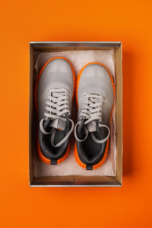 Pair of new gray mesh fabric sneakers in the open box on the bright orange background. Modern textile trainers with grooved orange sole for sport and active lifestyle. Open new shoes box. Top view.の写真素材