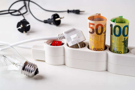 Fifty and one hundred euro banknotes plugged into the extension socket, few electric plugs and light bulb over white desk. Increasing of electricity cost, expensive energy, rise in electricity prices concepts.の写真素材