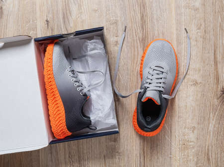 One sneaker in the open cardboard box and other near on the floor. Pair of new gray textile sneakers with grooved orange sole. Stylish mesh fabric trainers for active lifestyle. New shoes box top view.の写真素材