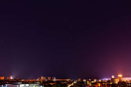 The constellation Orion shines brightly in the dark sky over illuminated city skyline at midnight. Clear starry sky over Varna in autumn. Night sky with bright stars over tranquil urban scene.の写真素材