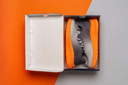 Pair of gray mesh fabric sneakers with grooved orange sole in the open box against orange gray...