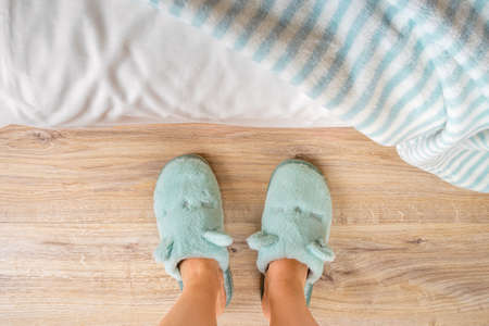 Female feet in soft blue slippers on a wooden floor near the bed close-up. Concepts of cozy fleece home shoes and warm domestic footwear for cold season. Top view.の写真素材