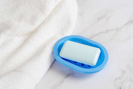 Light blue soap bar on a dish near white cotton terry towel over marble surface. Washing hands with...