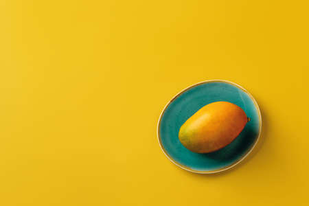 Whole ripe yellow Palmer mango on the turquoise plate over bright yellow background. Ready to eat...
