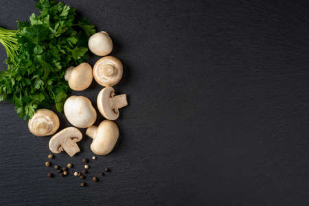 Raw champignons wole and halved and bunch of parsley over black slate slab. Cooking fresh button mushrooms Agaricus bisporus. Vegetarian menu. Recipe with vegetable protein. Copy space. Top view.の写真素材