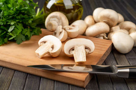 Raw champignon cut in half and and kitchen knife on a cutting board, whole button mushrooms and bunch of parsley over dark background. Vegetarian food ingredient, vegetable protein. Front view.の写真素材