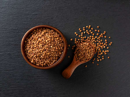 Wooden bowl and spoon full of raw organic buckwheat groats over black slate background. Ingredient...