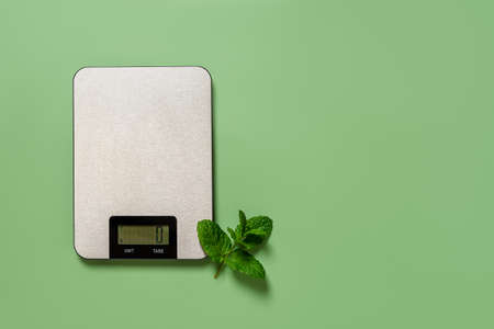 Digital kitchen scales and fresh mint sprig on a green background with copy space. Household measuring device for dieting and cooking. Electronic scales for food weight measurement. Top view.の写真素材