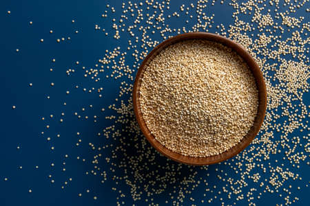Dry quinoa in a brown wooden bowl over blue background with scattered grains. White quinoa seeds for gluten free dieting. Superfood chenopodium quinoa. Vegetarian healthy eating. Copy space. Top view.の写真素材