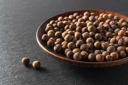 Allspice jamaica pepper on a wooden plate over black slate background. Whole dried berries of the Pimenta dioica tropical tree. Organic spices and seasonings concepts. Macro. Front view.の写真素材