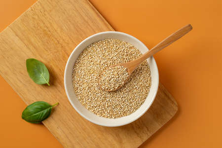 White quinoa grains and spoon in a beige bowl on a wooden cutting board over orange background. Chenopodium quinoa seeds for gluten free dieting. Concepts of superfood vegetarian healthy eating. Top view.の写真素材