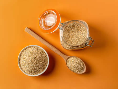 Top view of raw quinoa in a bowl, wooden spoon and clip top glass jar over bright orange background....