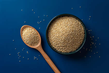 Raw quinoa grains in a wooden bowl and spoon over blue background. White quinoa seeds for vegetarian healthy eating. Cooking chenopodium quinoa concept. Gluten free superfood. Top view.の写真素材
